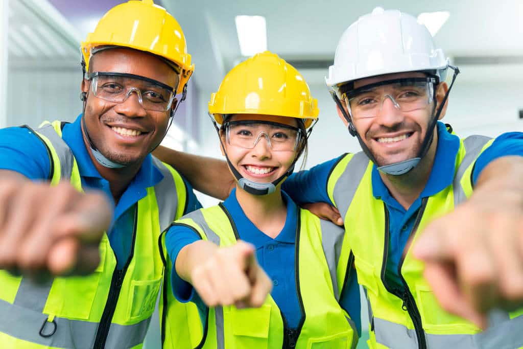 Três trabalhadores com capacete, óculos de segurança e colete amarelo de segurando apontando para câmera e sorrindo felizes, simbolizando a SIPAT