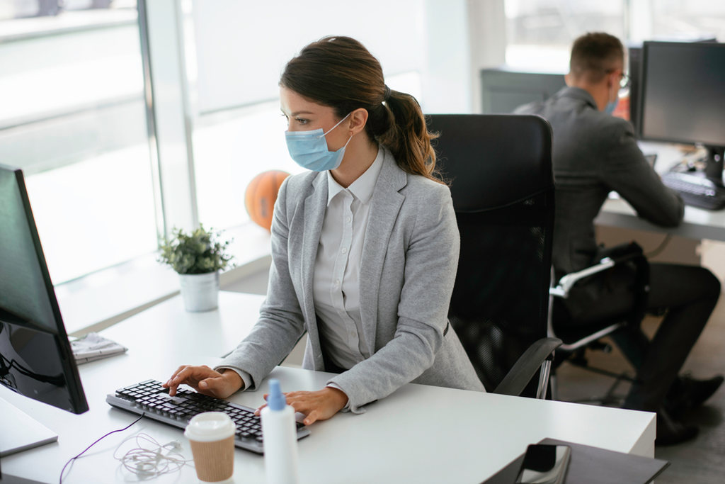 Mulher de máscara sentada em cadeira de escritório usando computador no trabalho, representando ergonomia e saúde