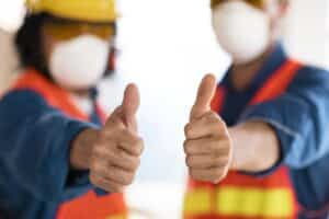 Dois profissionais fazendo sinal de jóia para a câmera e usando EPIs como capacete, máscara e colete; simbolizando por que investir em segurança do trabalho